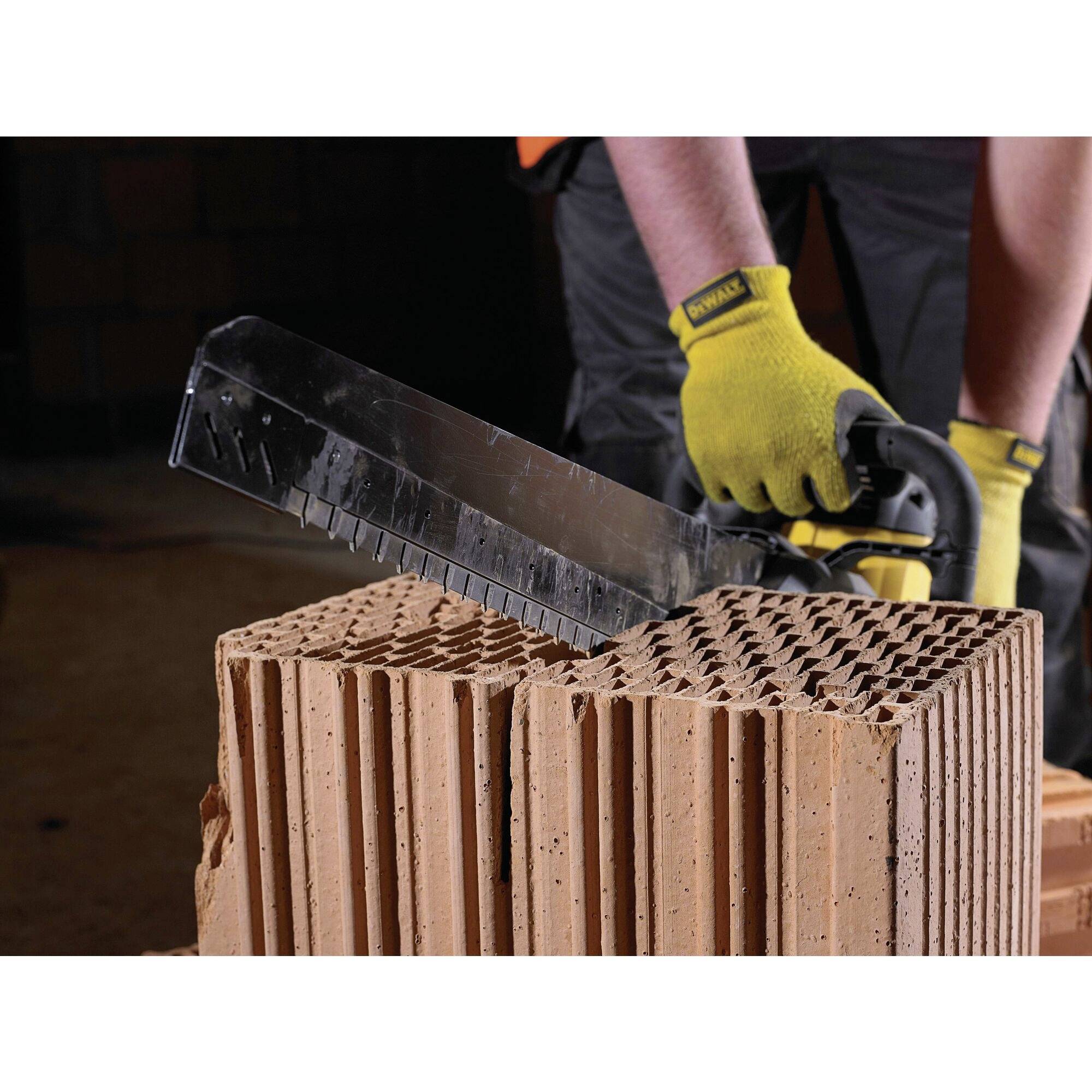 A person wearing yellow gloves is sawing a large, hollow brick with a handsaw. The brick is positioned on a table.