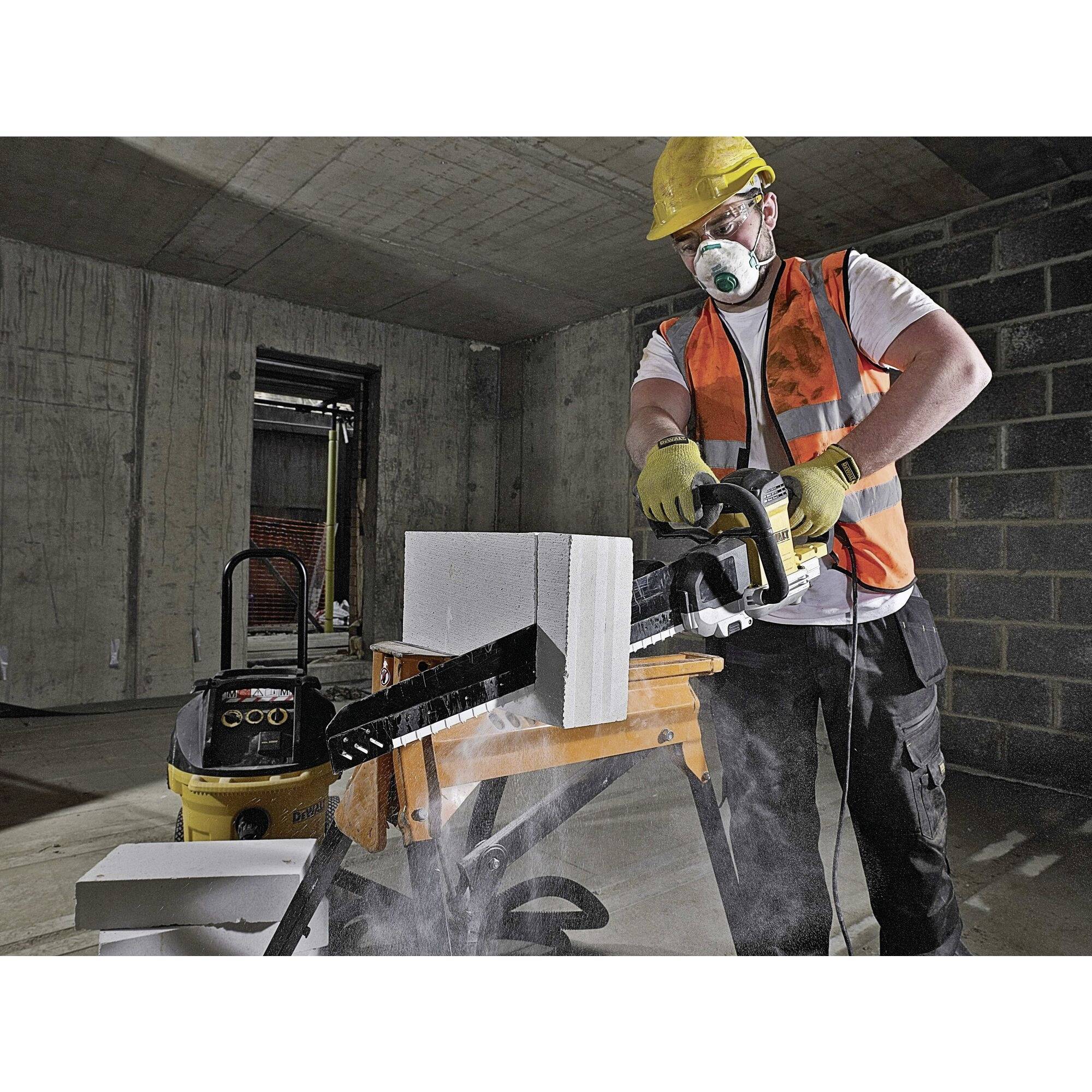 A construction worker in safety clothing is cutting concrete blocks with a saw in a partially completed room. Dust is rising.