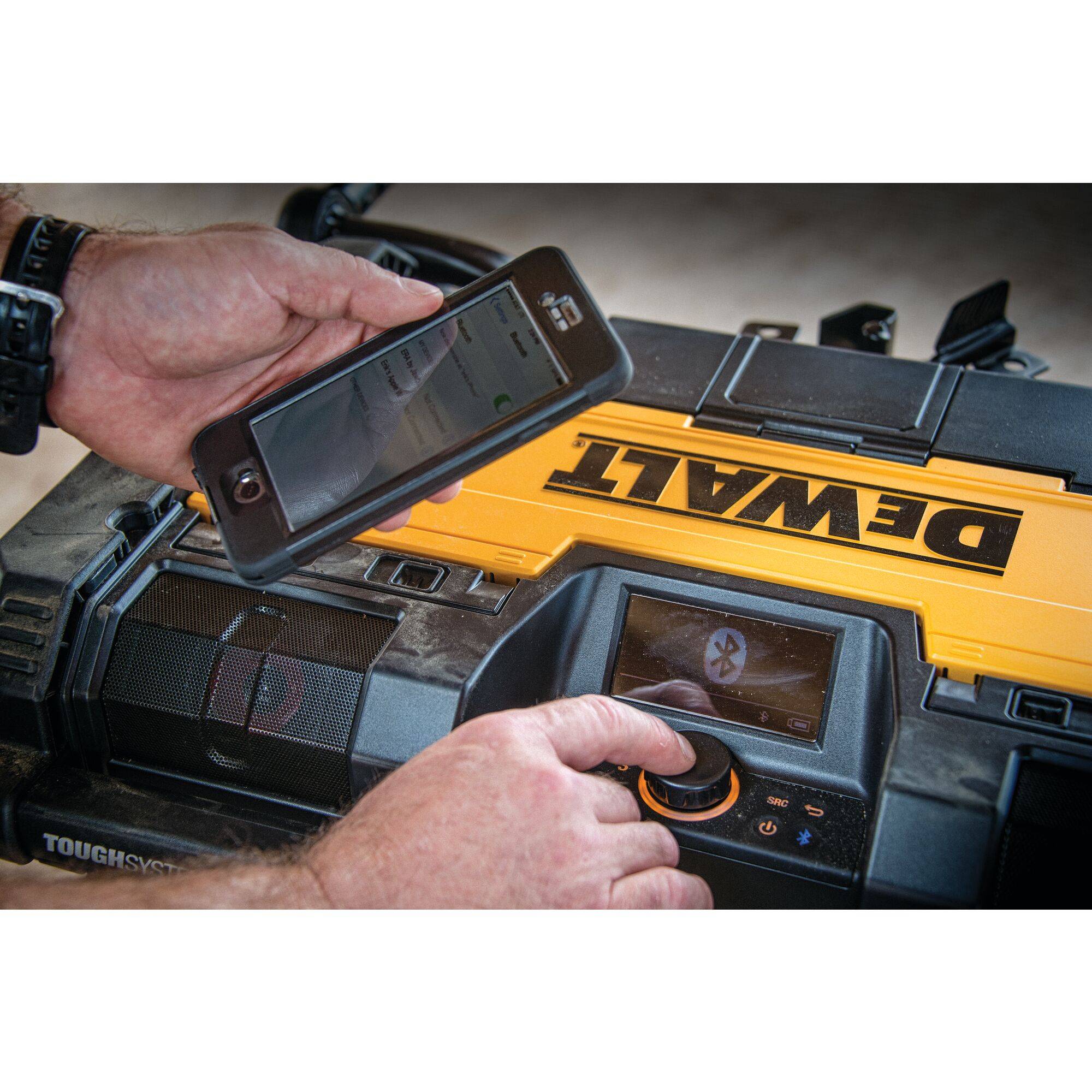 A person is operating a smartphone connected to a yellow DEWALT music system. The display shows a Bluetooth connection.