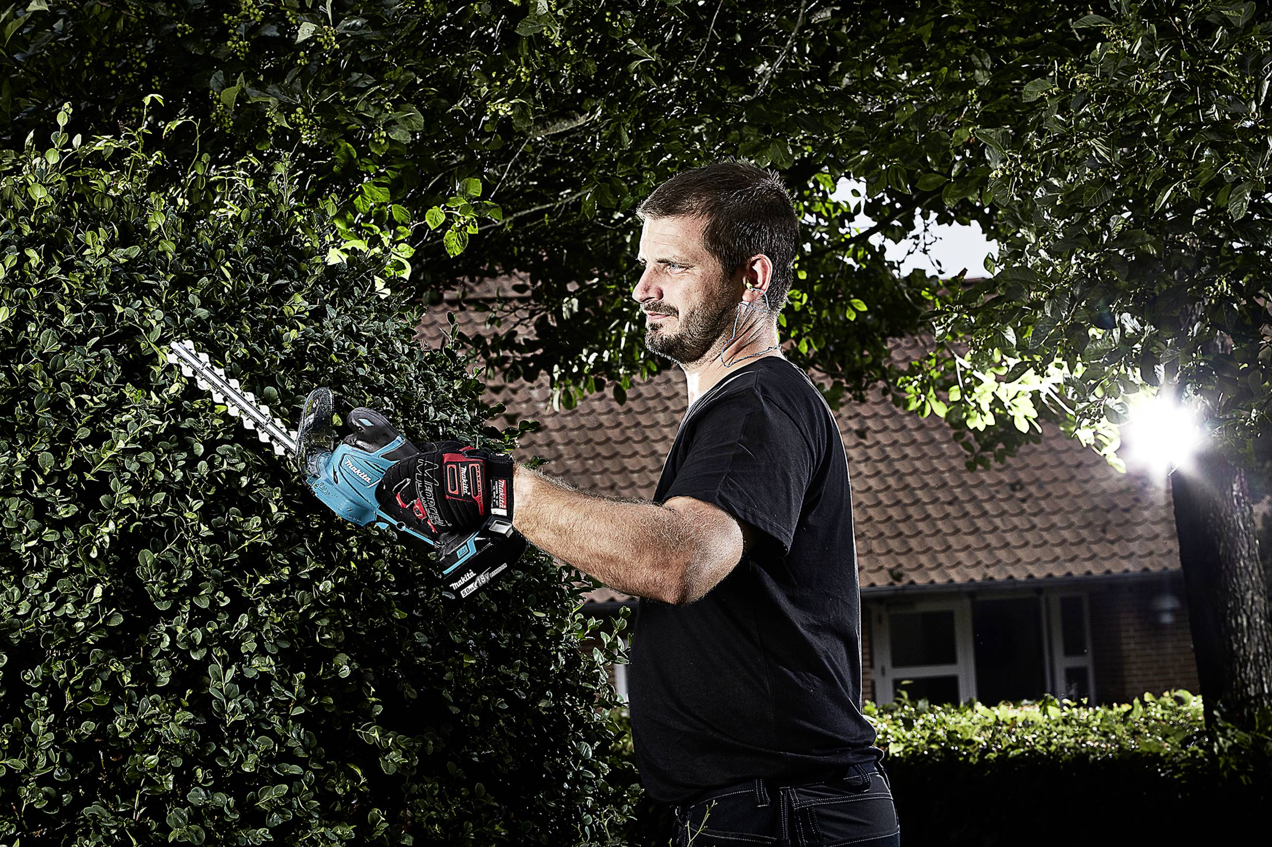 A man is trimming a green hedge with an electric hedge trimmer, surrounded by trees. A brick building is visible in the background.