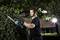A man is trimming a green hedge with an electric hedge trimmer, surrounded by trees. A brick building is visible in the background.