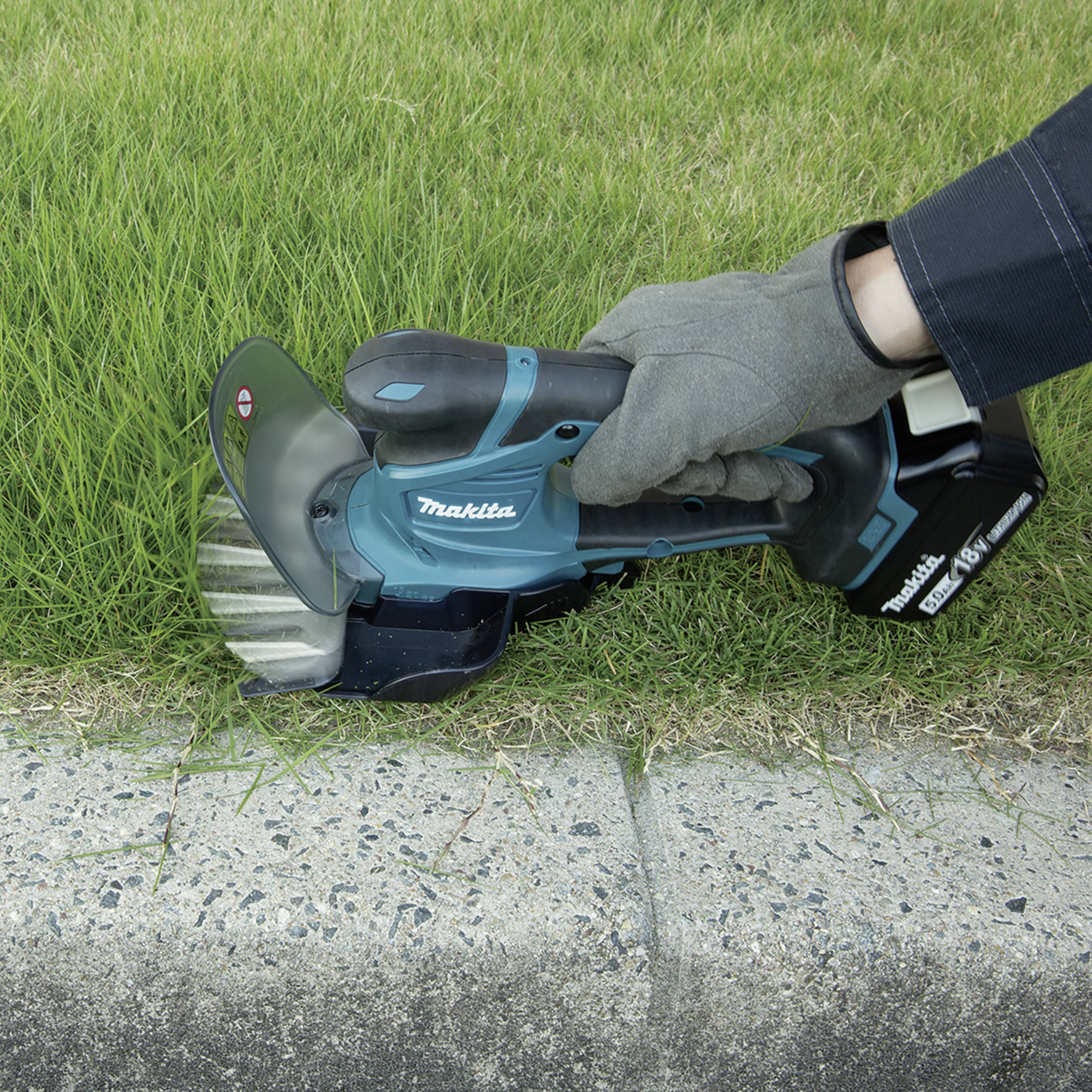 A person is trimming the grass along a pavement using a cordless strimmer.