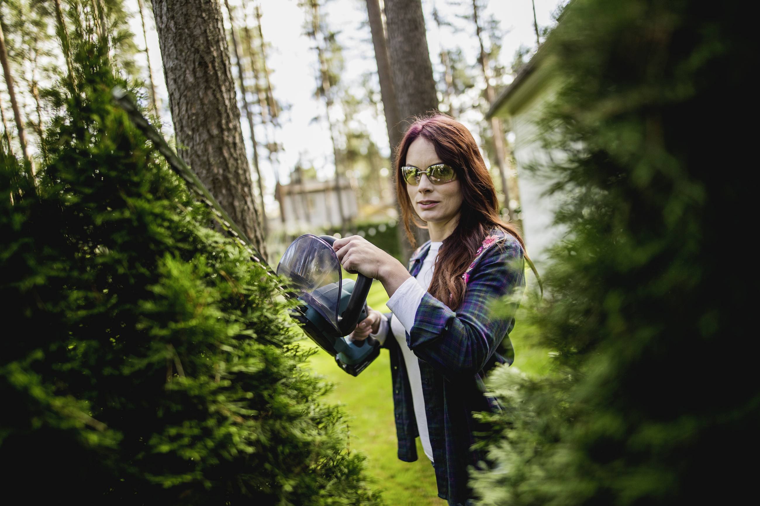 A woman wearing sunglasses is trimming bushes outside with hedge shears.