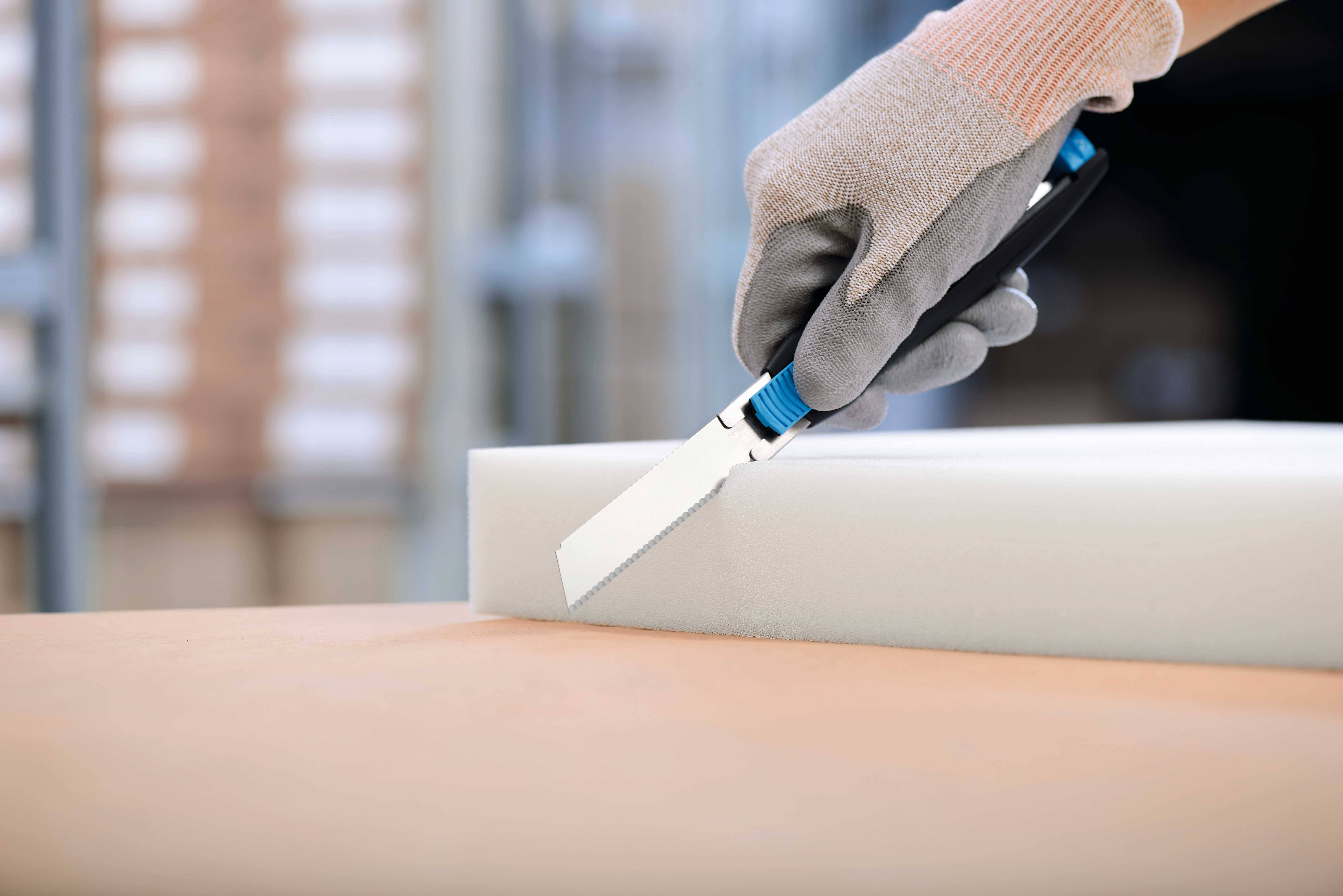 A hand wearing a glove is cutting through a piece of foam with a craft knife on a table.