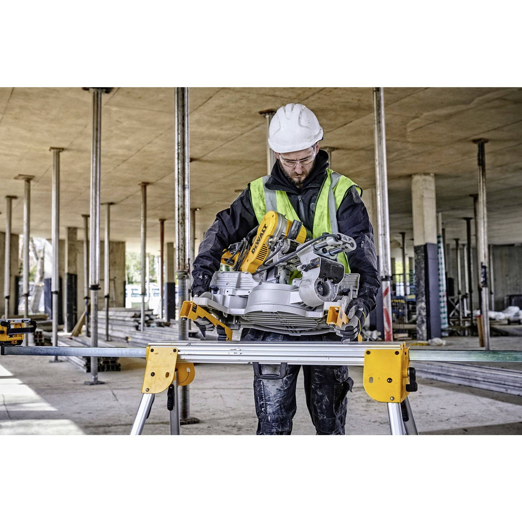 Worker wearing a hard hat and high-visibility vest operating a saw on a construction site, surrounded by concrete supports and building equipment.