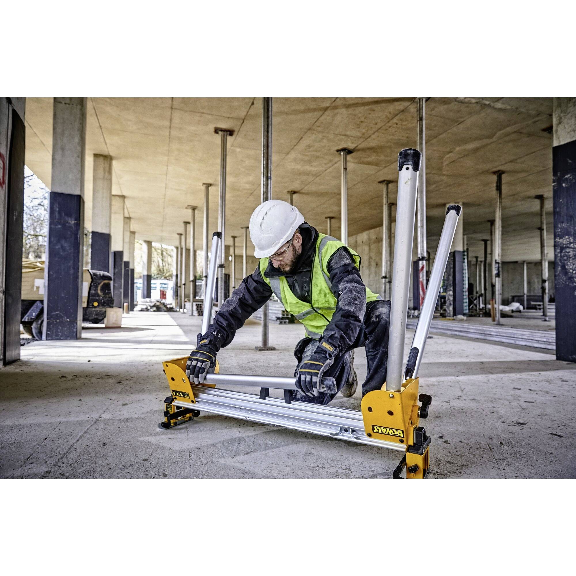 A construction worker in protective clothing and hard hat adjusts a building support in an unfinished building. The background shows columns and construction equipment.