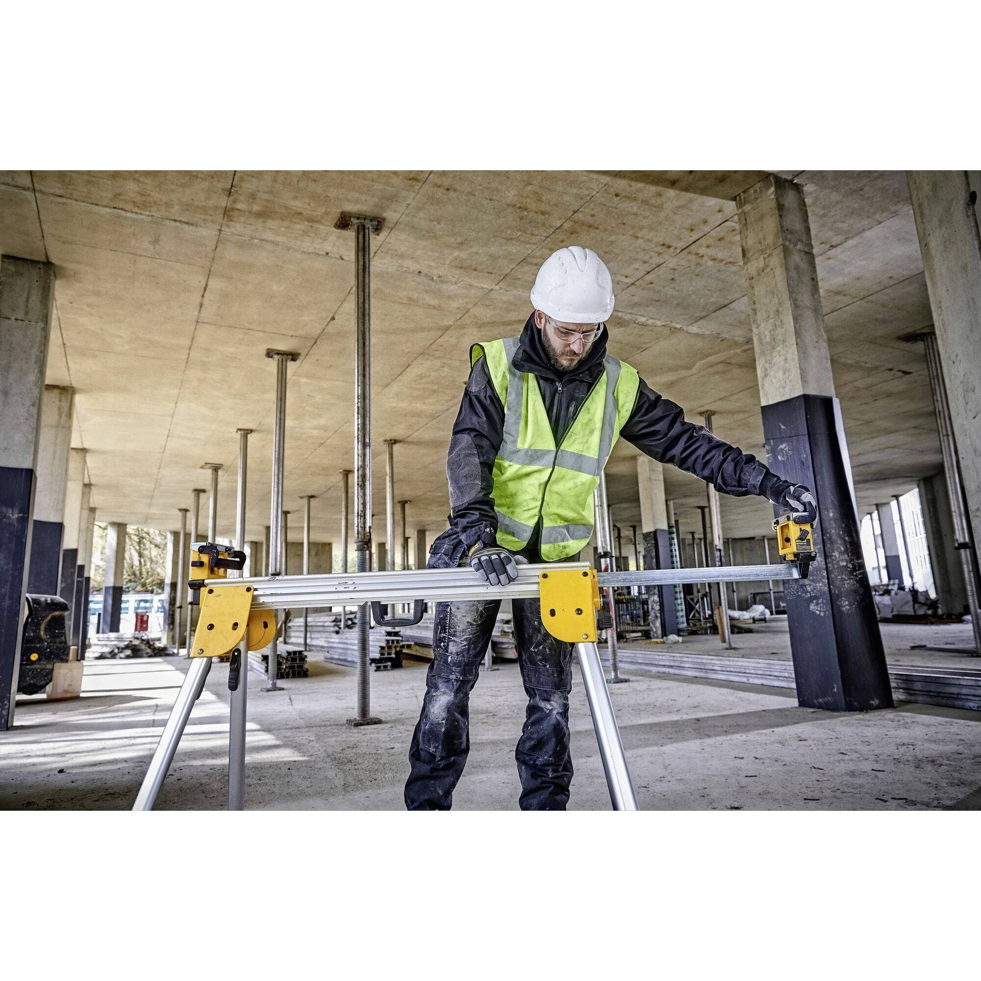 A construction worker wearing a hard hat and high-visibility vest is working in an unfinished building. He is measuring a long metal piece on a tripod.