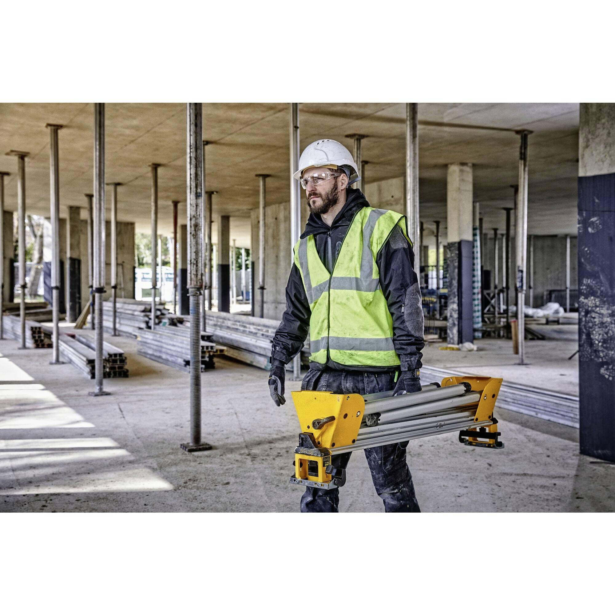 A construction worker in a high-visibility vest and hard hat is carrying a yellow ladder through a concrete structure on a building site.