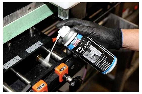 A gloved hand sprays lubricant from a can onto a metal machine part, likely for maintenance or reducing friction.