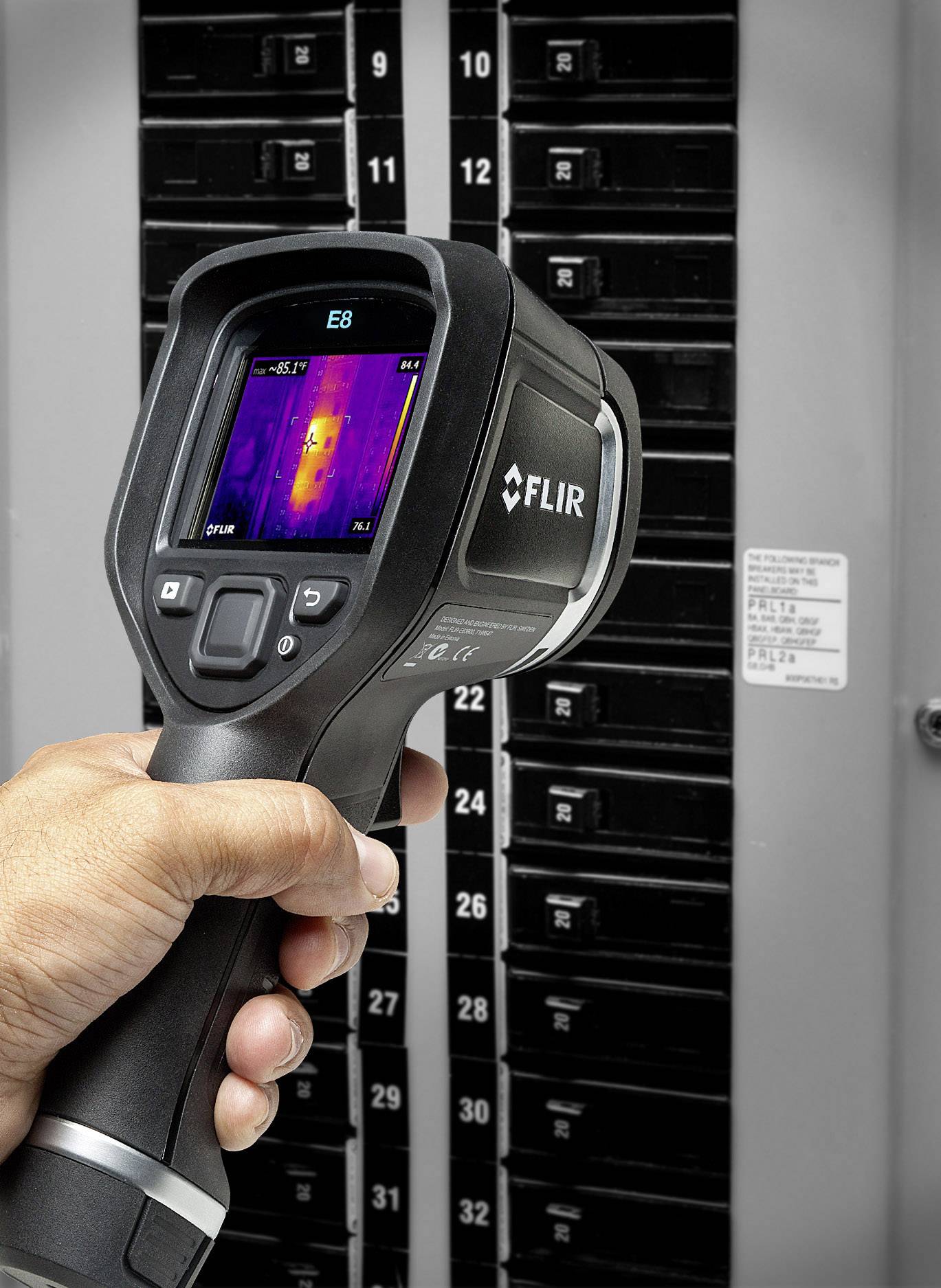 A person is holding a thermal imaging camera in front of an open fuse box. The screen displays a thermal image of the box.