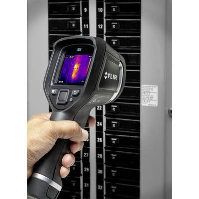 A person is holding a thermal imaging camera in front of an open fuse box. The screen displays a thermal image of the box.