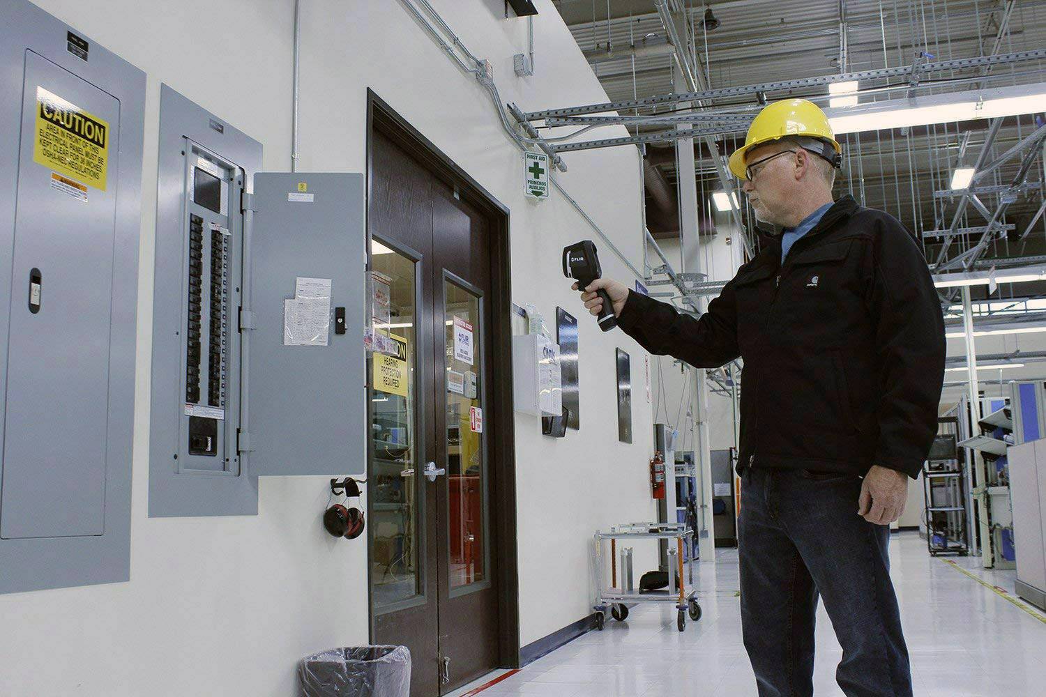 A person wearing a hard hat and safety glasses is using an infrared thermometer to inspect an electrical control panel in a factory hall.