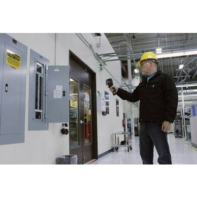 A person wearing a hard hat and safety glasses is using an infrared thermometer to inspect an electrical control panel in a factory hall.