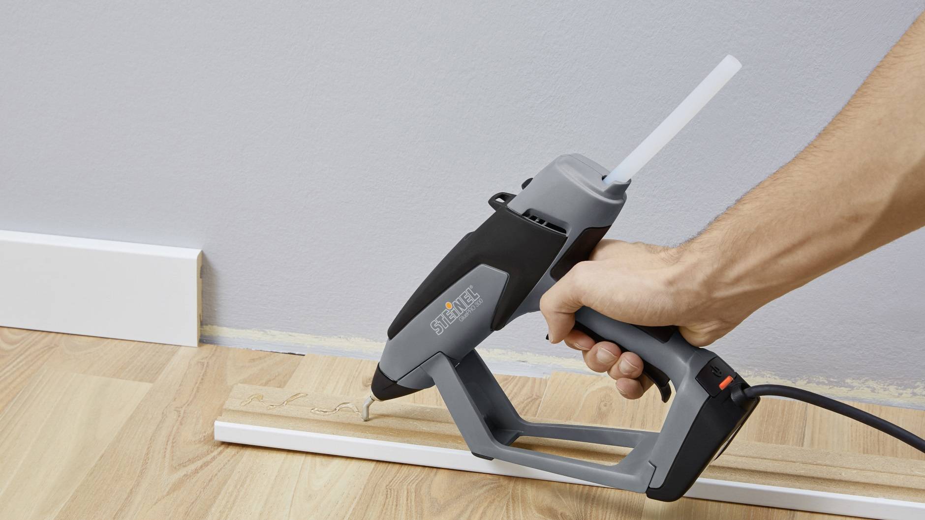 A person is using a hot glue gun to attach a piece of wood to a floorboard in front of a wall.