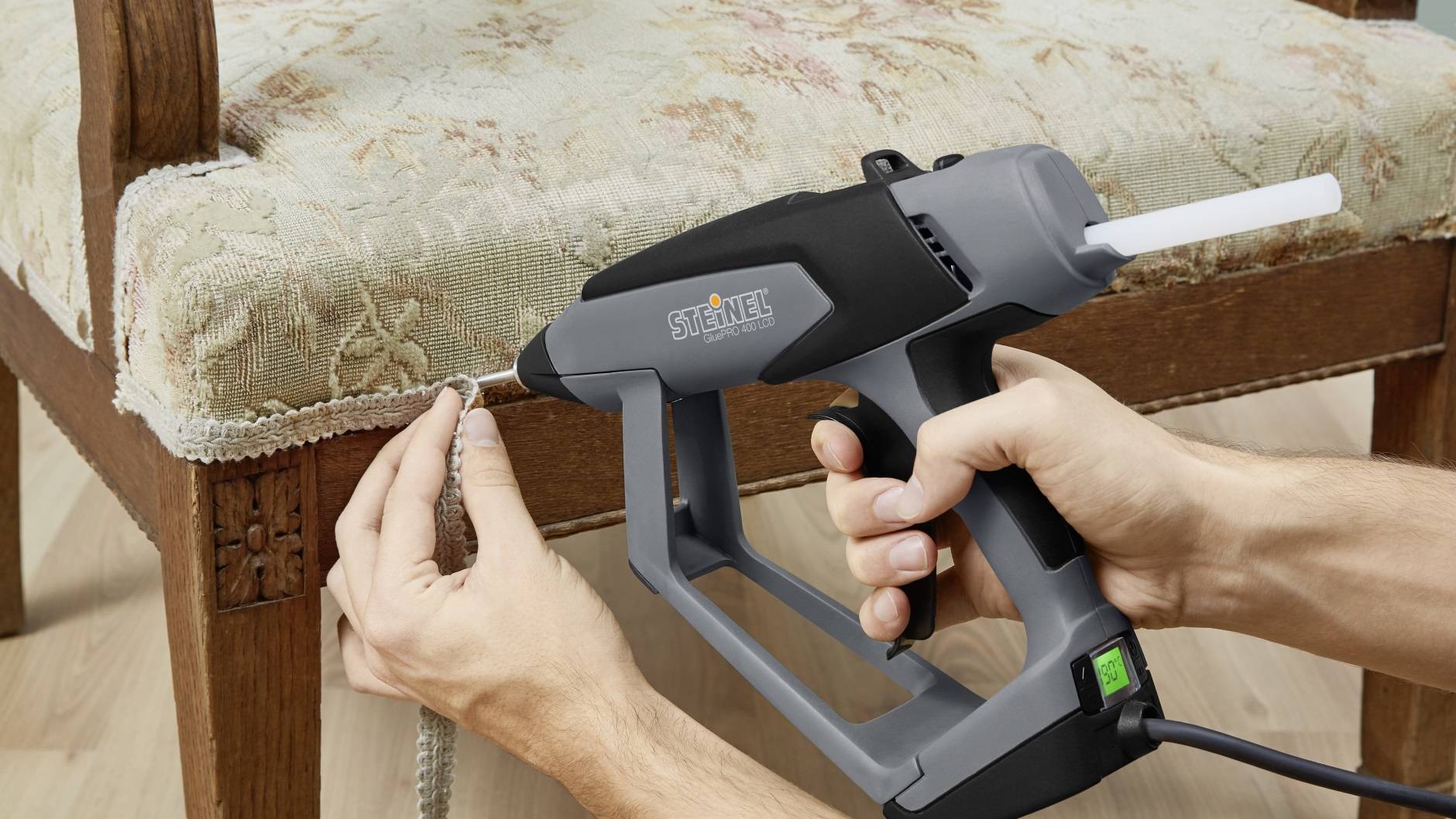 A person is attaching a decorative trim to an antique upholstered chair using a hot glue gun.