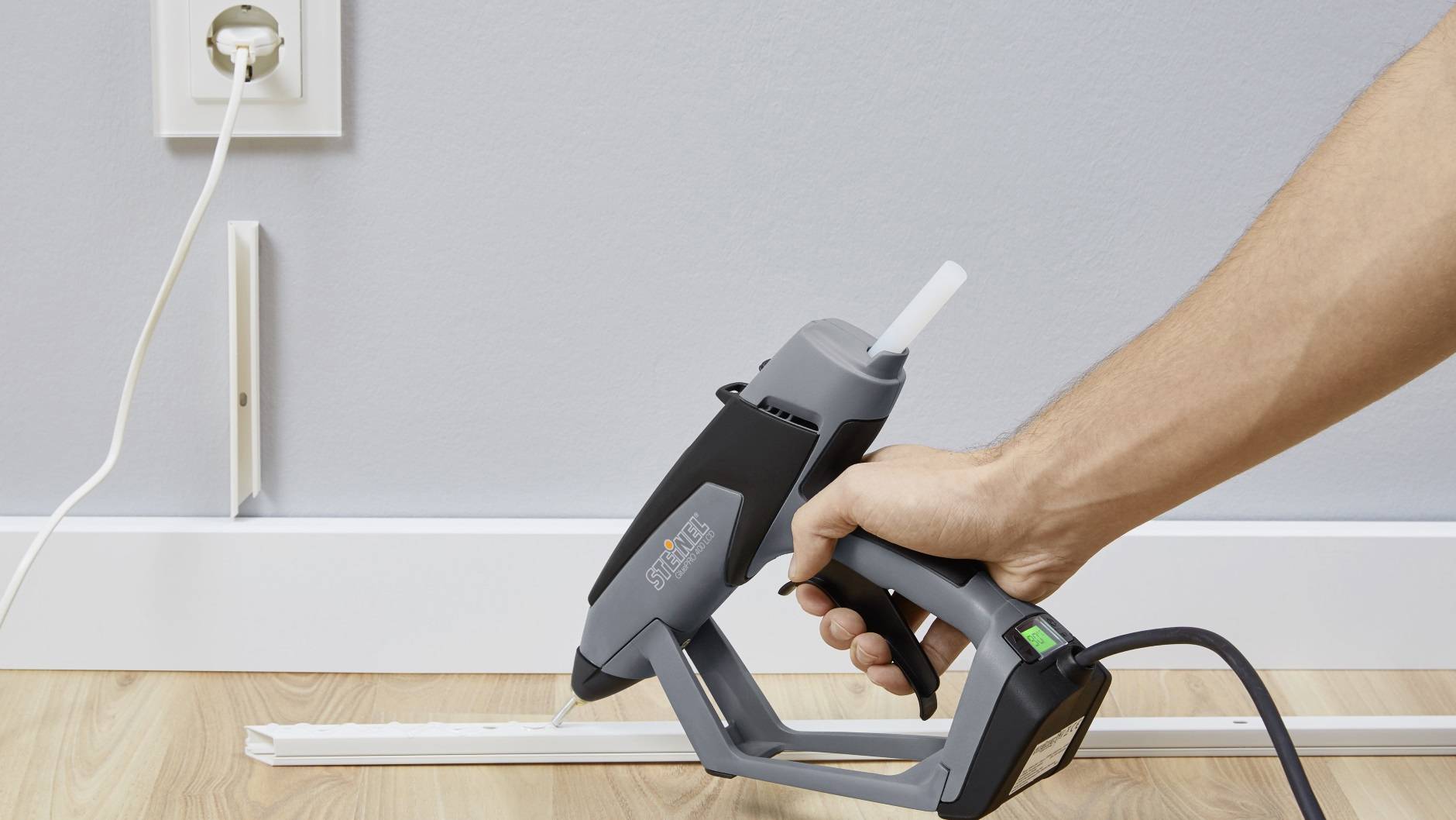 A person is holding a hot glue gun and securing a cable cover along a skirting board. A power socket is visible in the background.
