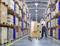 A warehouse worker pushes a trolley laden with boxes through a large, well-organised warehouse with high shelves stacked with additional boxes.