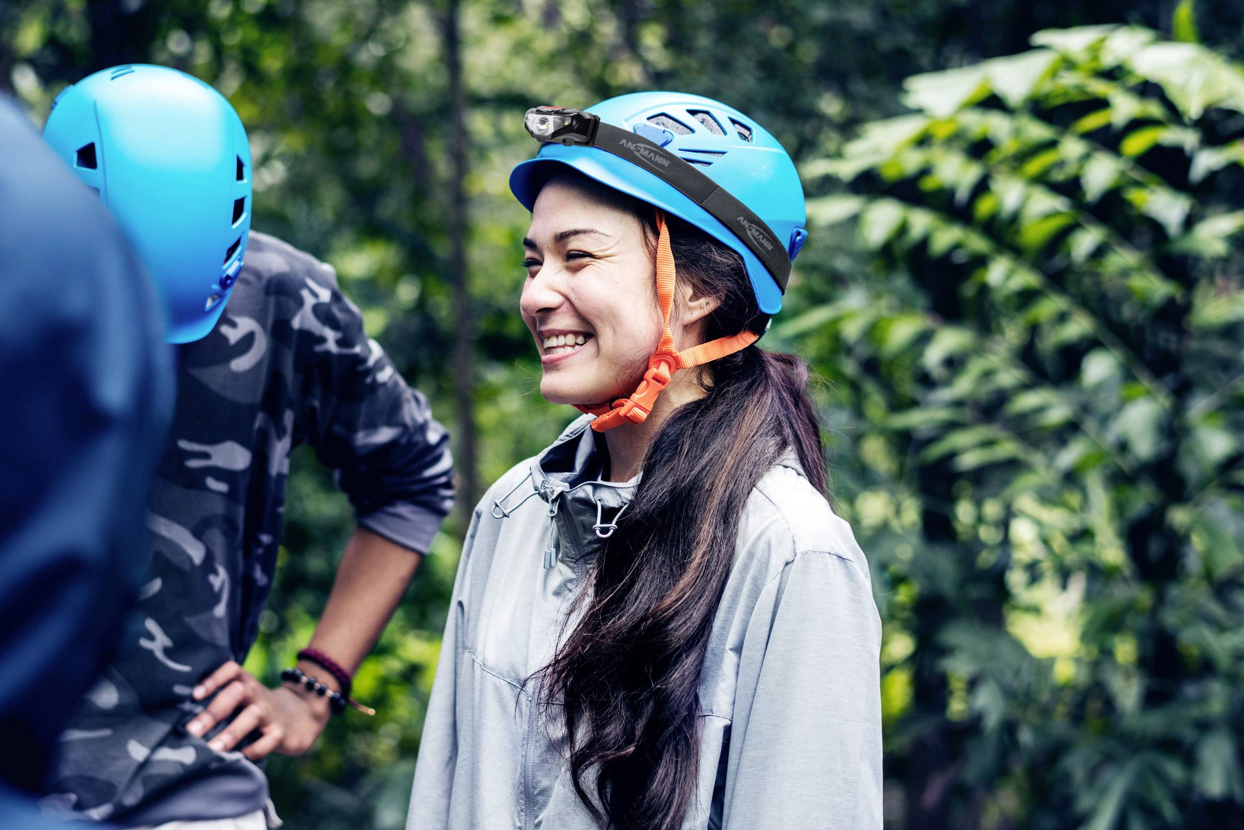 A smiling person is wearing a blue climbing helmet and standing in a wooded area, surrounded by green vegetation.
