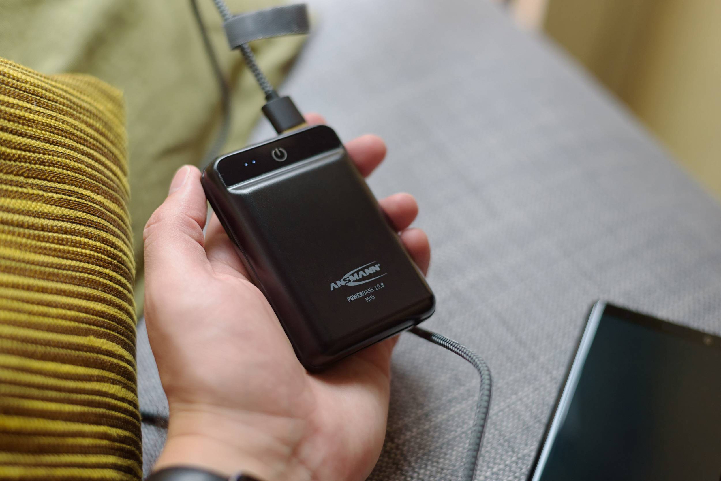 A person is holding a black, portable power bank connected to a charging cable. A sofa is visible in the background.