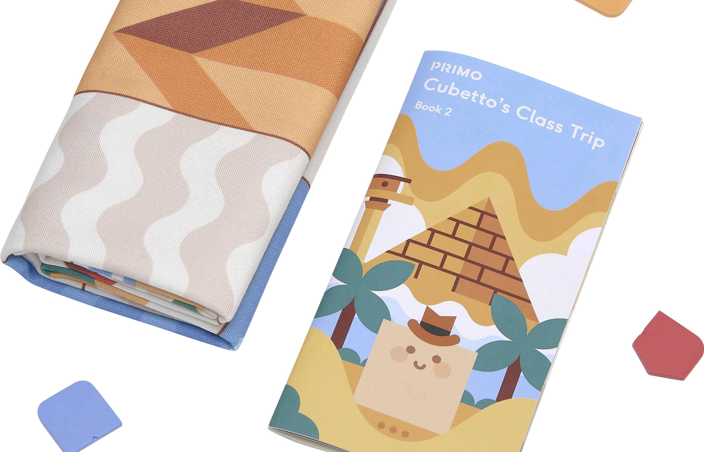 Book titled 'Cubetto's Class Trip', illustrated cover featuring a pyramid, palm trees, and a cube-shaped figure. Beside it, a fabric with patterns.