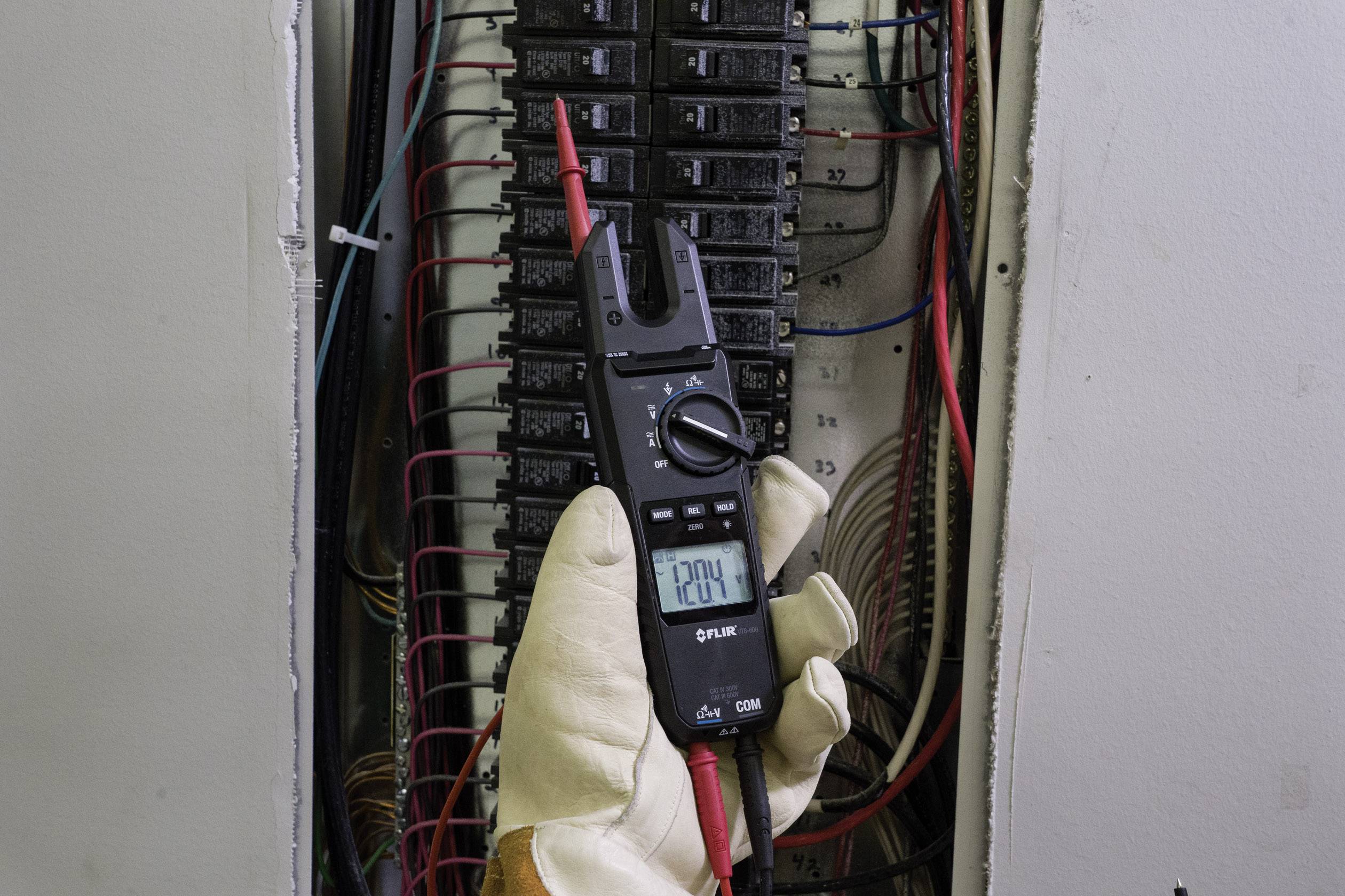 A person is measuring the voltage in an electrical cabinet with a multimeter, displayed value: 120.4 V. They are wearing safety gloves.