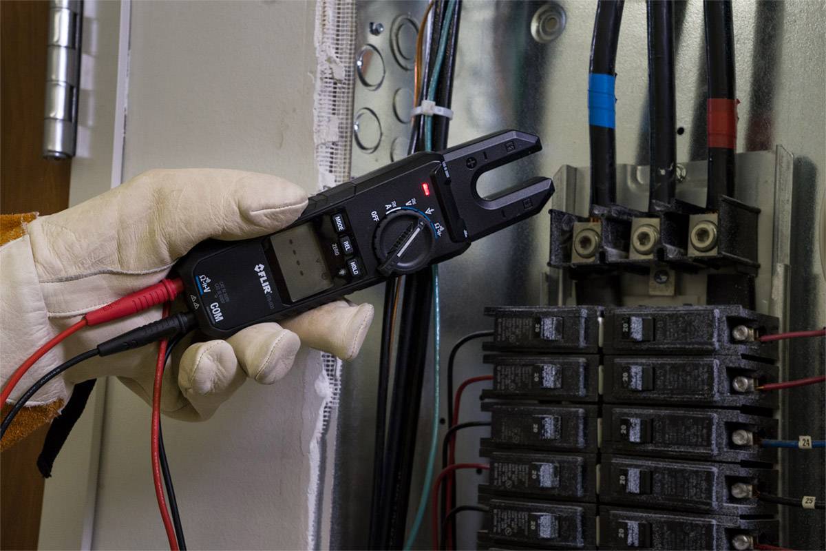A hand is holding an electrical measuring device in an open fuse box. The device is displaying a measurement, with cables and fuses visible.