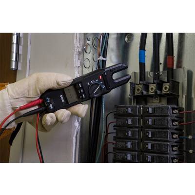A hand is holding an electrical measuring device in an open fuse box. The device is displaying a measurement, with cables and fuses visible.