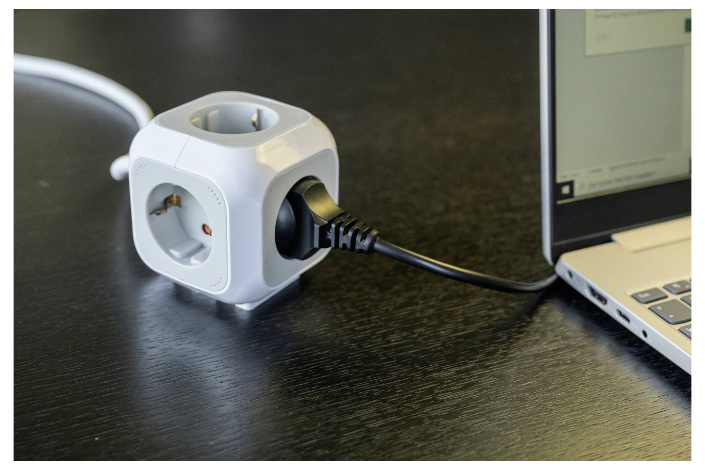 White cube power strip with multiple outlets and a plugged-in black cord beside a laptop on a desk.