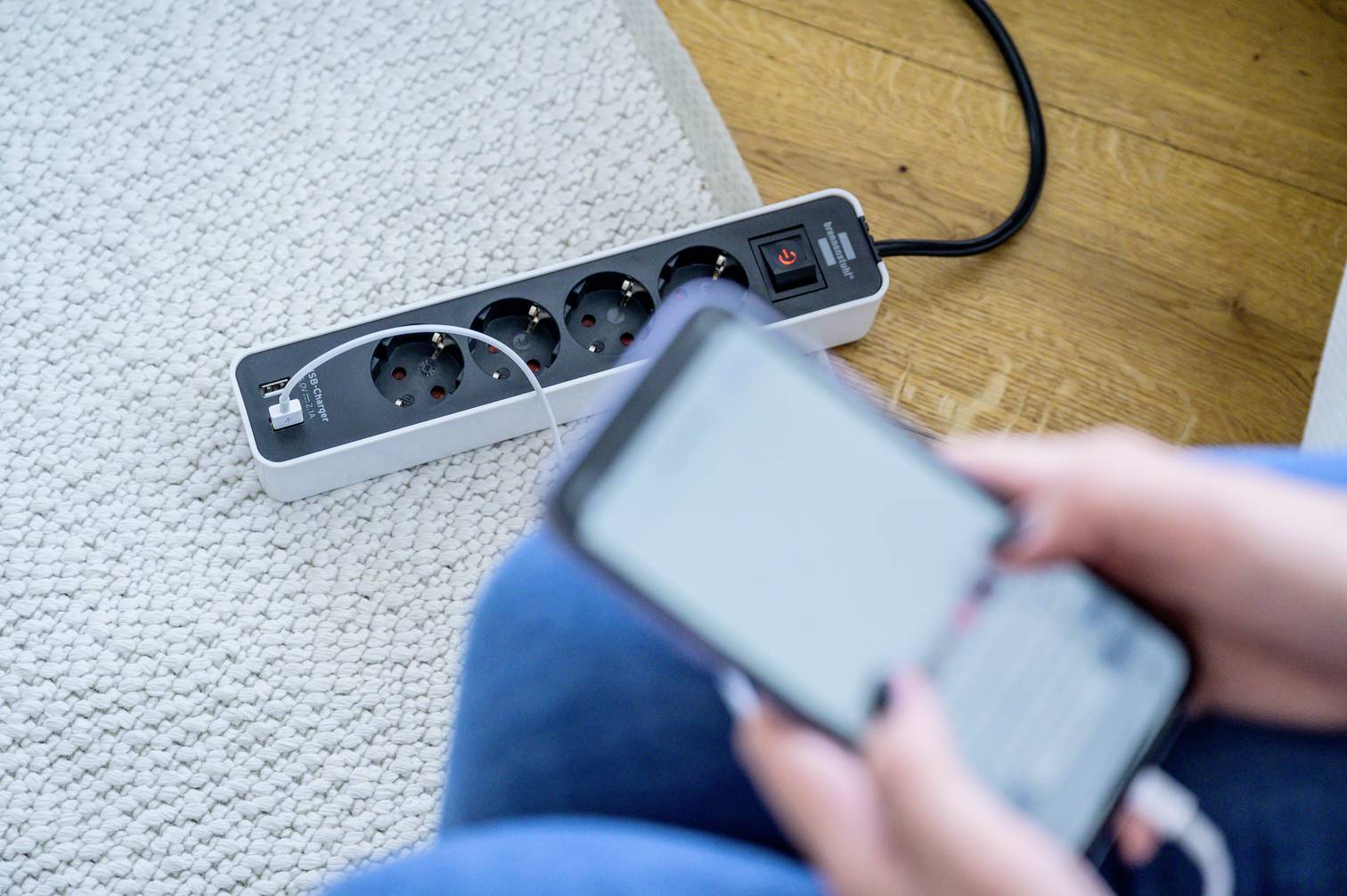 A person is holding a mobile phone; in the background, a power strip with a charging cable plugged in lies on the floor.