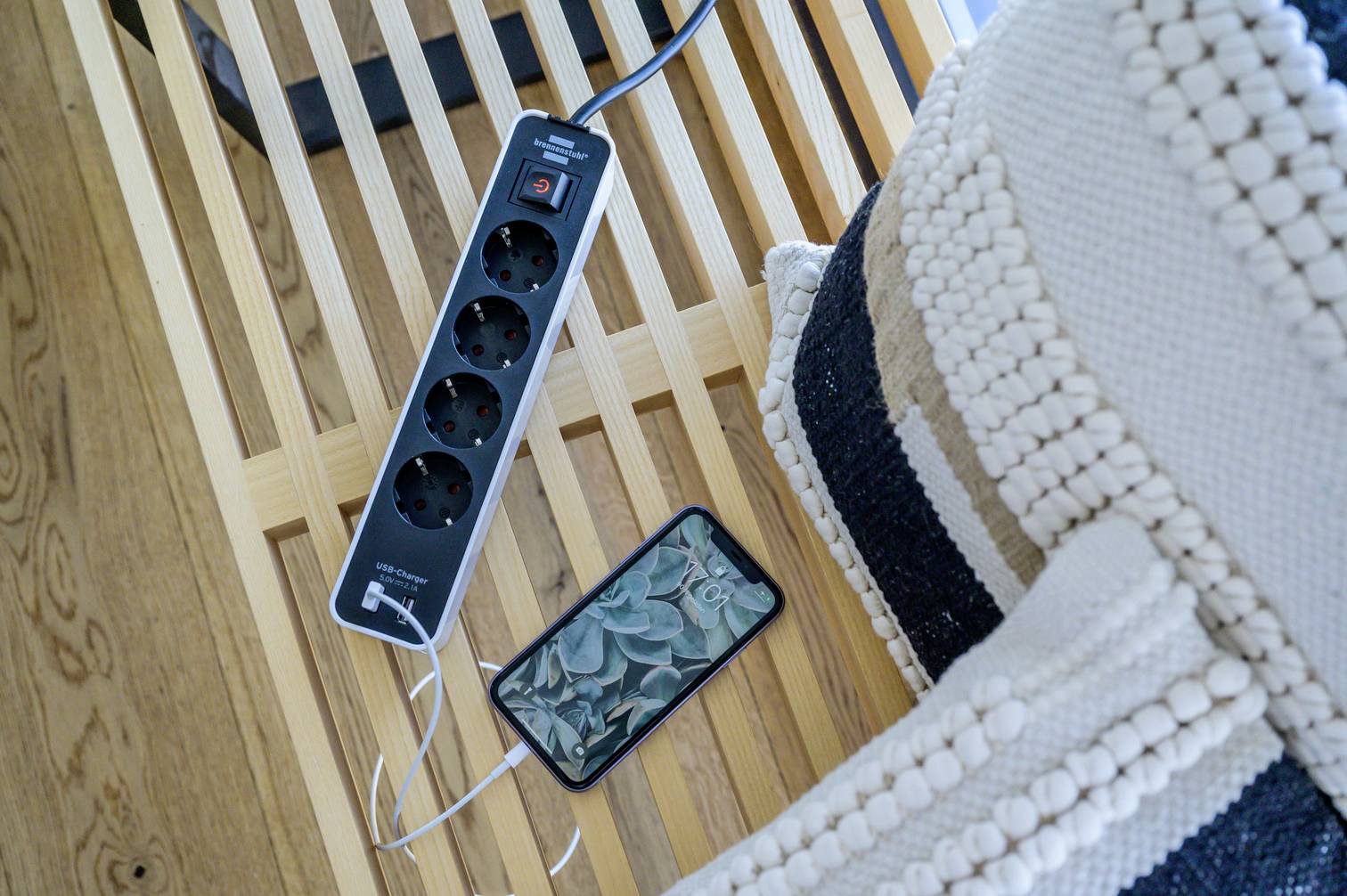 'A mobile phone is connected to a black multi-socket adaptor with a charging cable, which is lying on a wooden table next to cushions.'