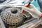 A hand wearing a glove cleans a bicycle cassette with a red tool. Background of metal parts, working in a bicycle workshop.