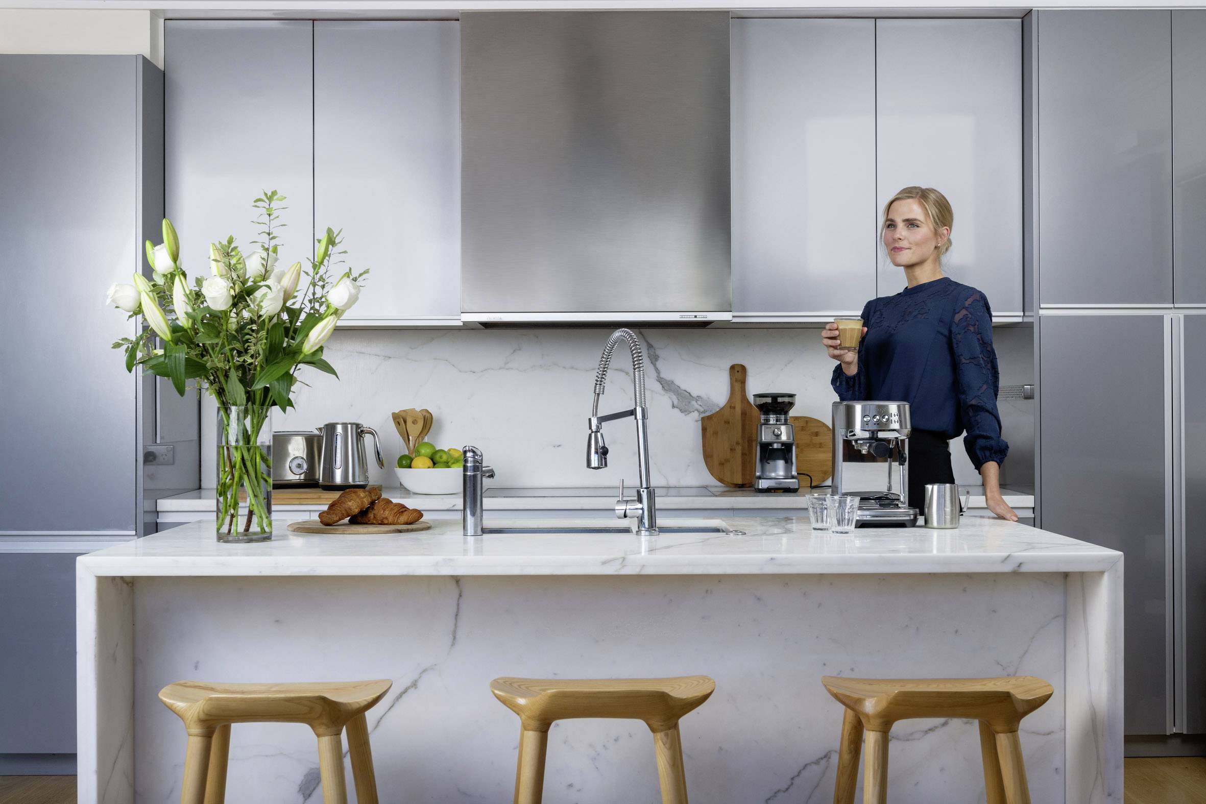 A woman stands at a modern kitchen island, holding a cup of coffee. Flowers, bread, and kitchen utensils are on the table.