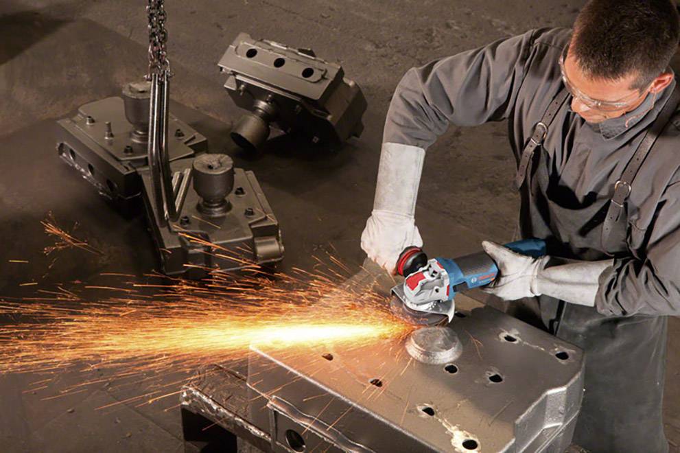 A worker is grinding a metal workpiece with an angle grinder. Sparks are flying. Further workpieces are visible in the background.