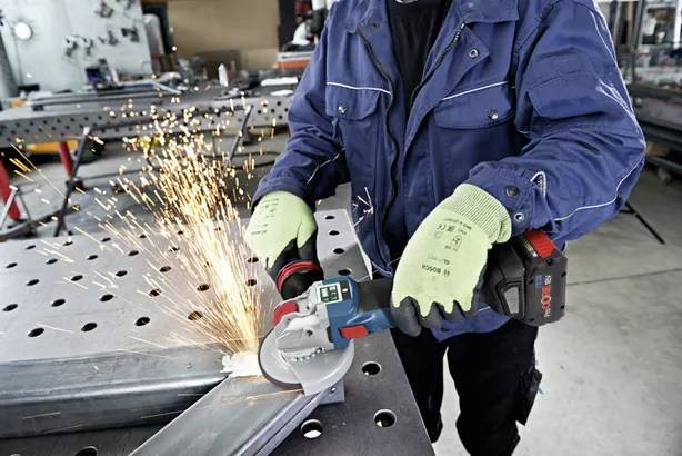 A person wearing workwear is grinding metal with an angle grinder, sparks flying. They are wearing green gloves and a blue jacket.