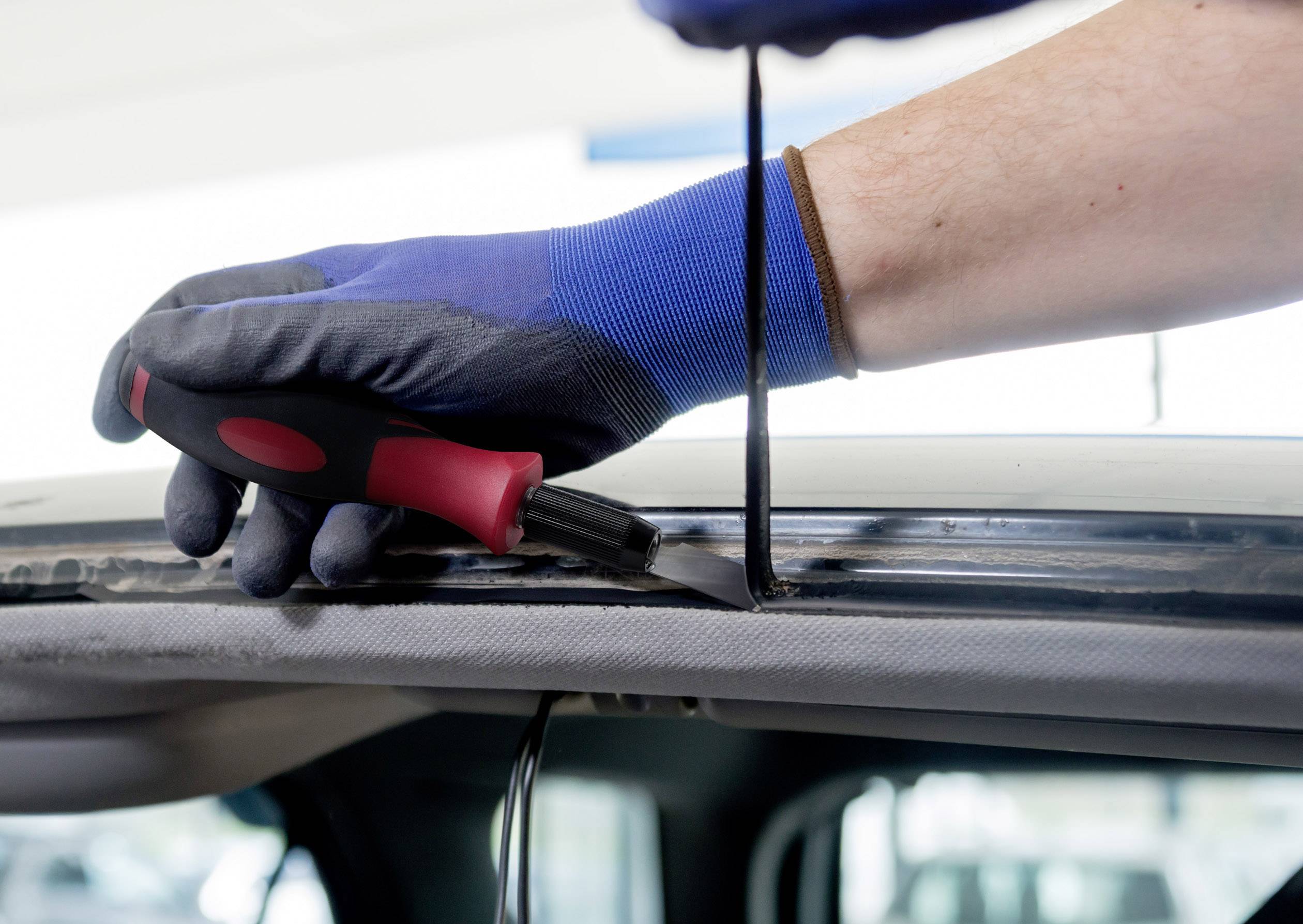 Someone is using a knife to remove the seal of a car window. The person is wearing blue work gloves.