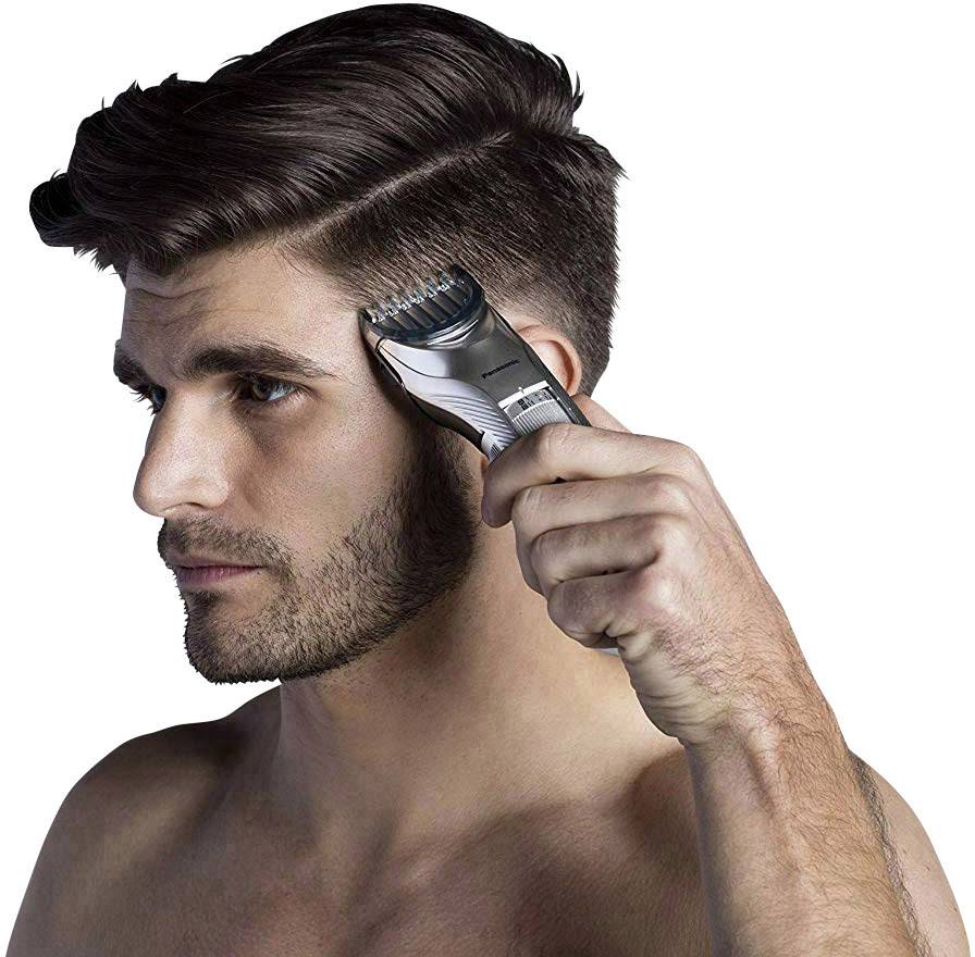 Man with beard trims hair using an electric hair clipper. Short sides, longer top hair. Focused on haircutting process.