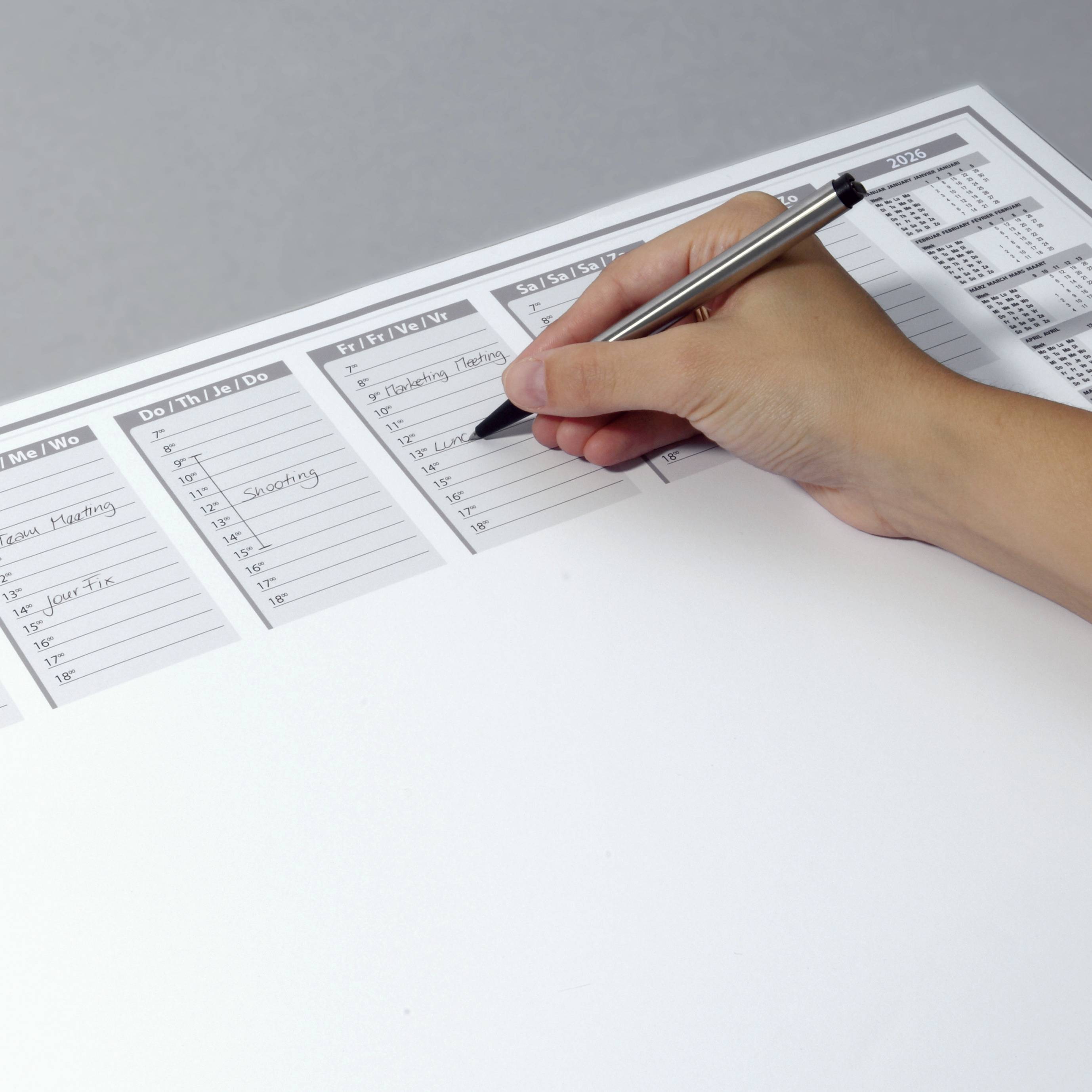 A hand is writing with a silver pen on a desk calendar displaying multiple columns of days and tasks.