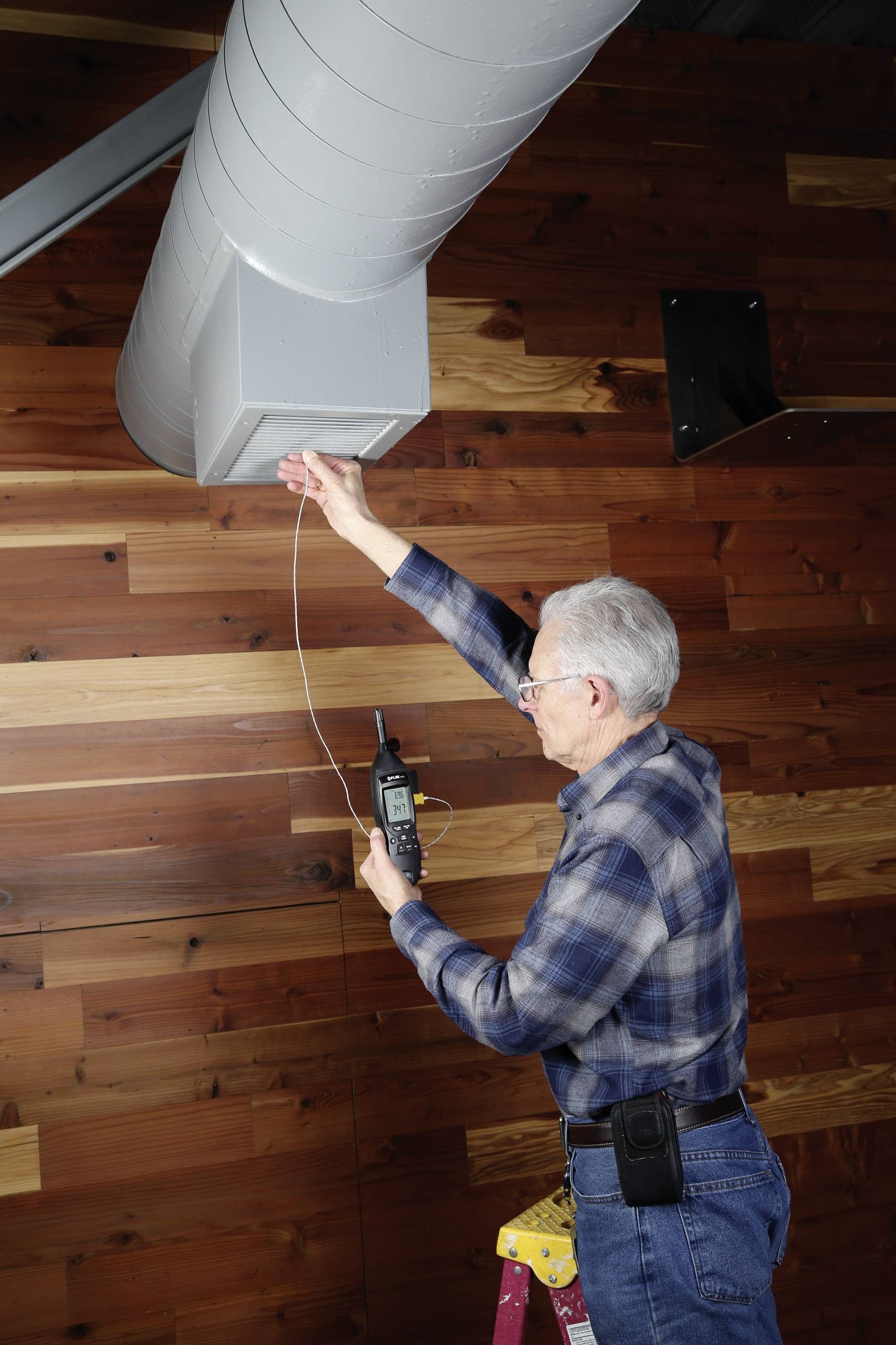 An older man in a checked shirt is measuring air quality or temperature using a device at a large ventilation system on the ceiling.