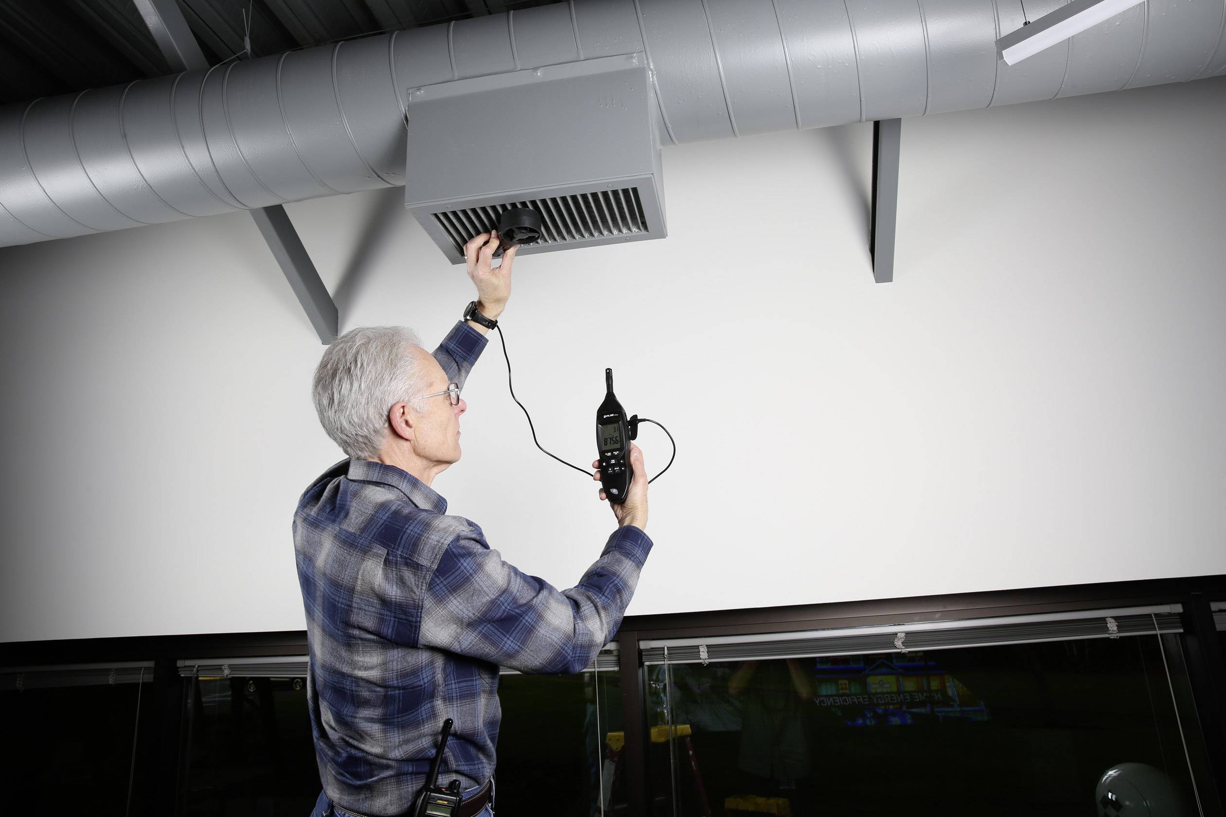 A person is inspecting a ventilation system, holding a measuring device to check air quality or air flow.