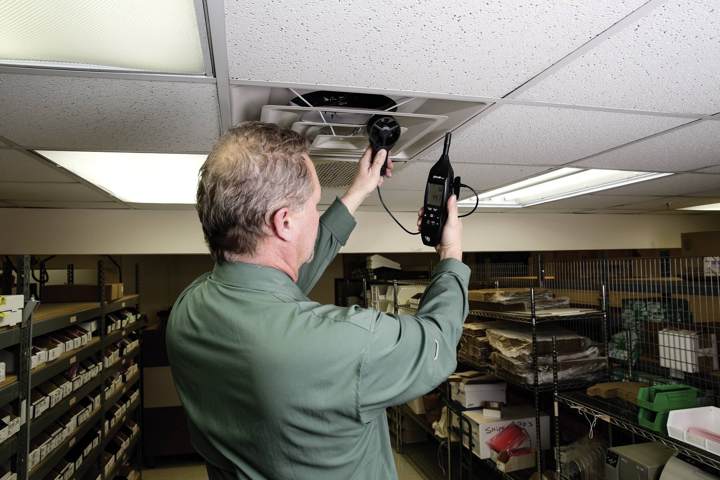 A man in green clothing is measuring air quality or temperature in a storage room using a handheld device while standing near the ceiling.
