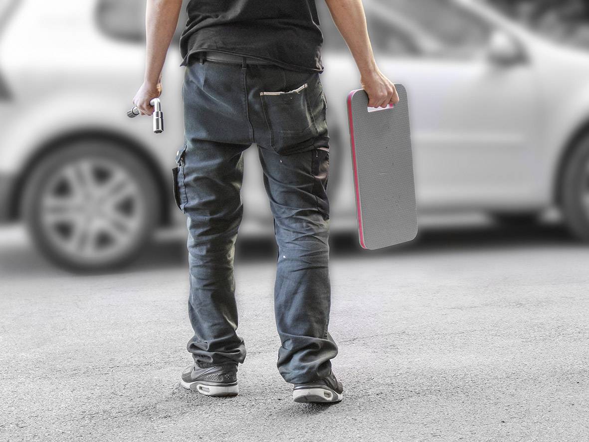 A worker stands on a street wearing work attire, holding a tool and a rectangular object, with a car in the background.