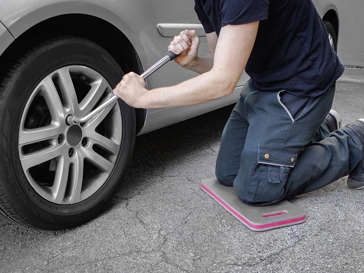 A person is kneeling on a mat and changing a car tyre with a spanner on a concrete surface.