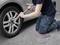 A person is kneeling on a mat and changing a car tyre with a spanner on a concrete surface.