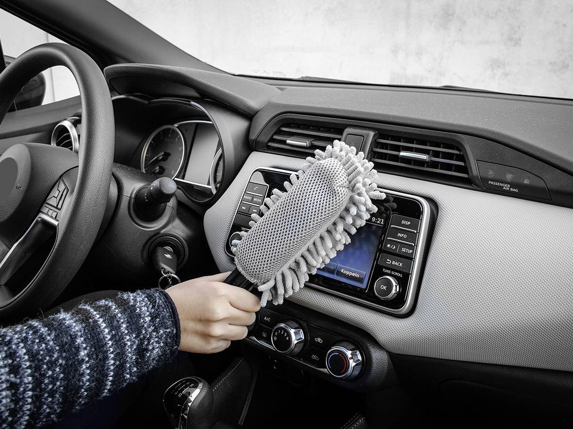 A person is cleaning the dashboard of a car with a grey feather duster. The display shows an audio system and control buttons.