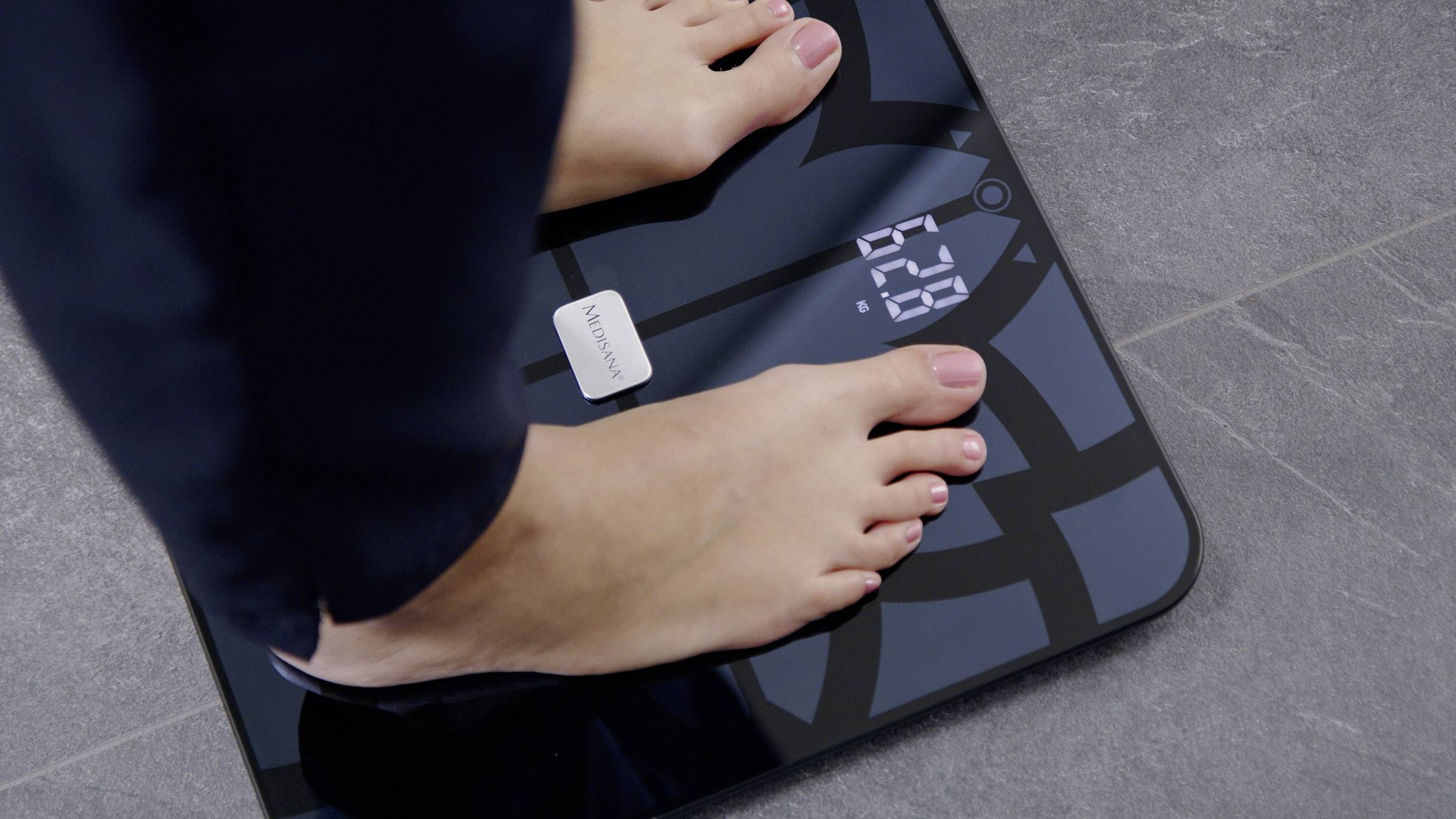 A person is standing on a digital scale, with the display showing '62.0'. The scale has a dark design with a geometric pattern on a grey floor.