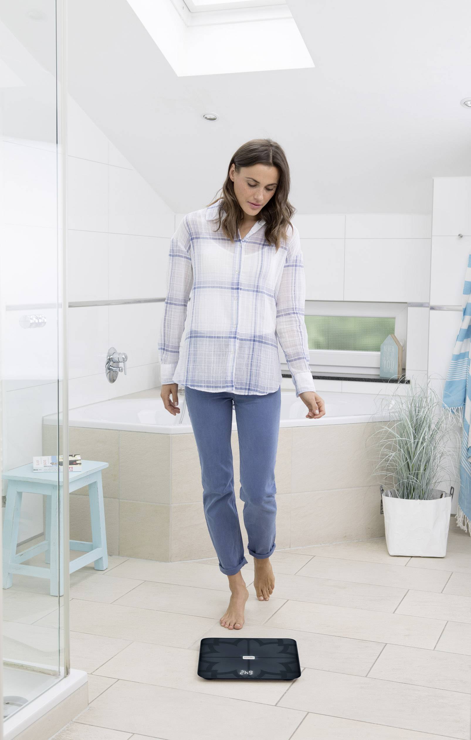 A woman is standing barefoot on a digital scale in a modern bathroom. She is wearing comfortable clothing and appears focused.