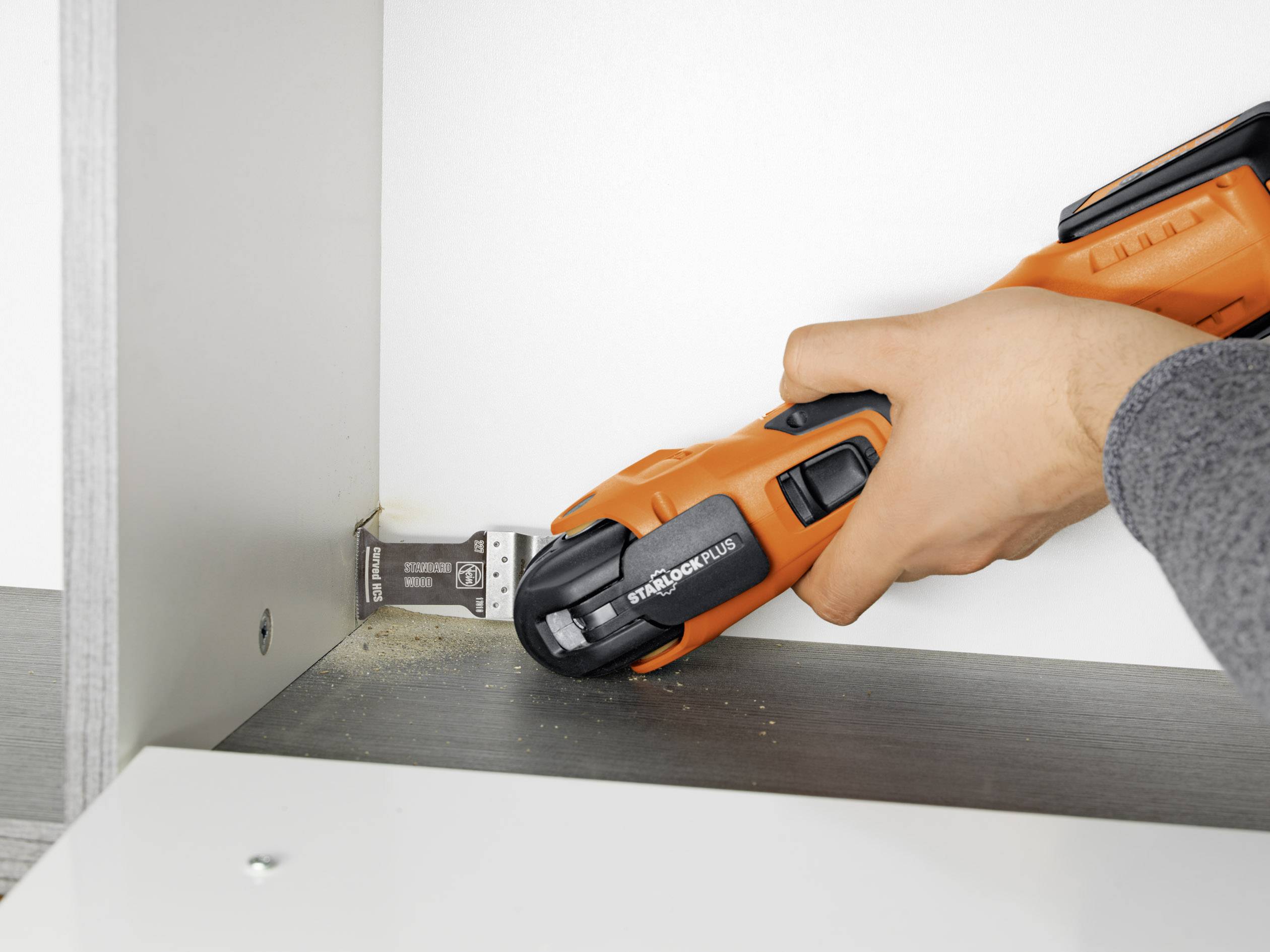 A hand is holding an oscillating multi-tool, sawing wood in a piece of furniture. Wood shavings are visible.