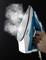 A steam iron is held upright while steam emerges from the openings. The appliance is designed in blue and white.