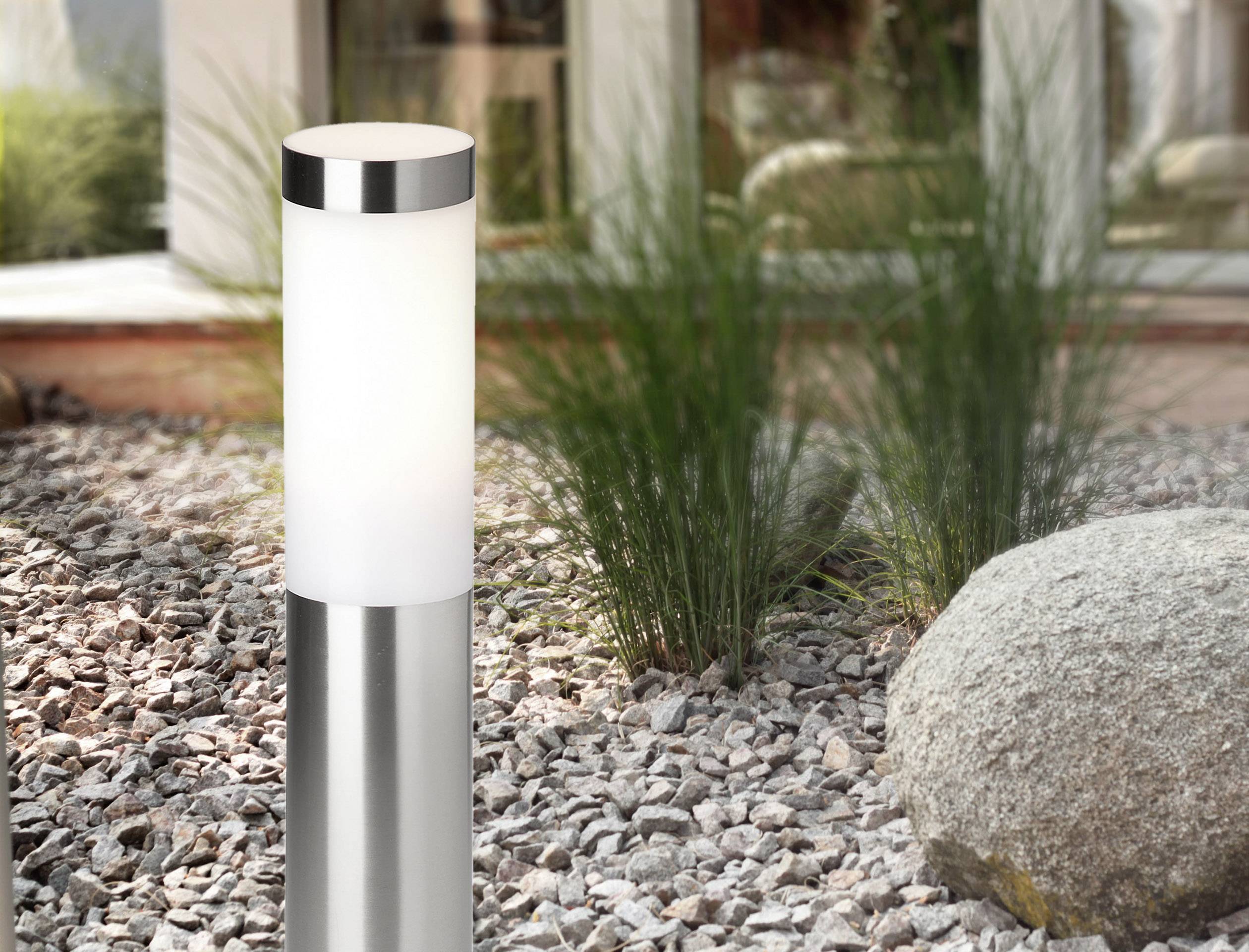 A modern outdoor light made of metal and glass stands in a gravel garden next to a large stone and grasses.