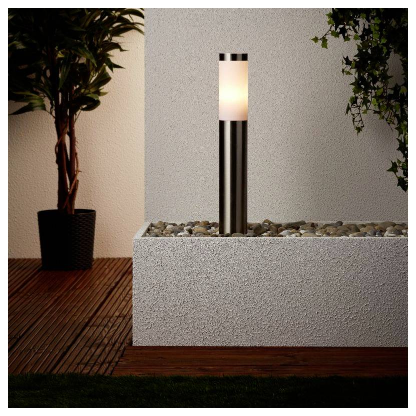 A modern outdoor lamp with a cylindrical metal base and soft white light on a pebble-filled platform, next to a potted plant.